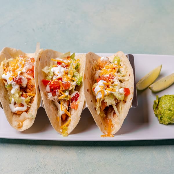 Three shrimp tacos with cheese, lettuce, tomato, and sour cream, with guacamole and lime wedges.