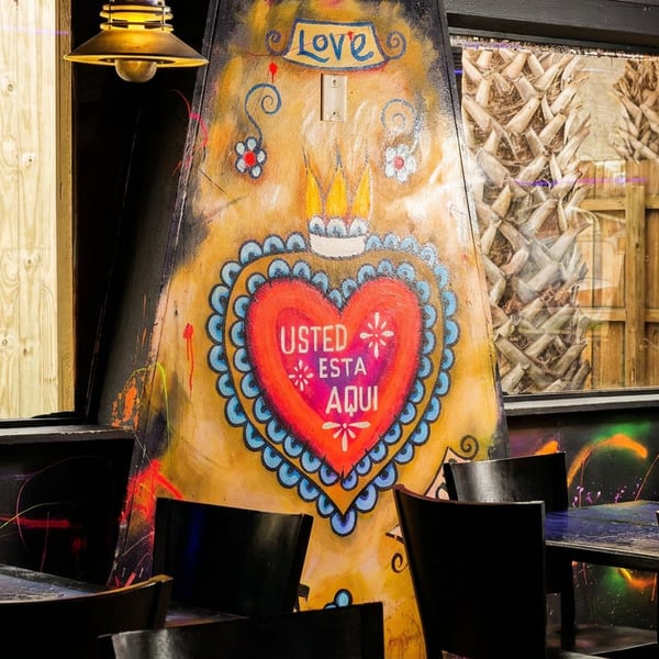 Restaurant interior with a large, colorful heart mural, dark tables, and chairs, with windows showing a wooden fence.