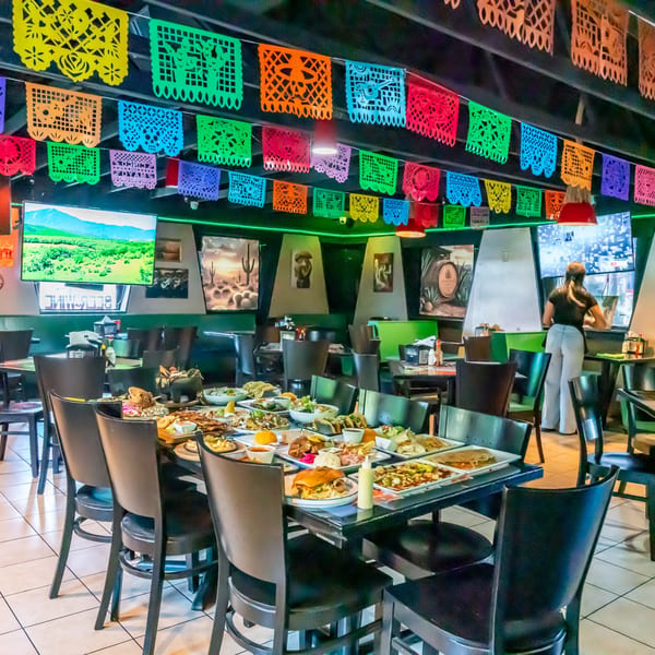 A vibrant Mexican restaurant interior with colorful papel picado banners, a long table laden with food, and a server.