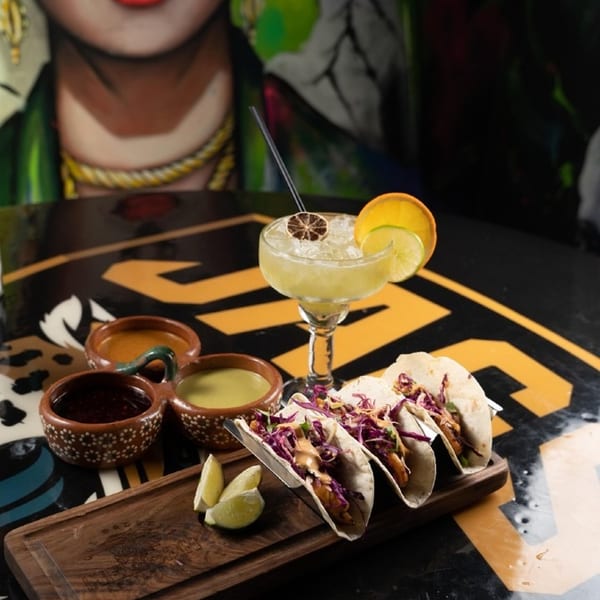 Three tacos, a margarita, and three dipping sauces on a dark table with a colorful mural.