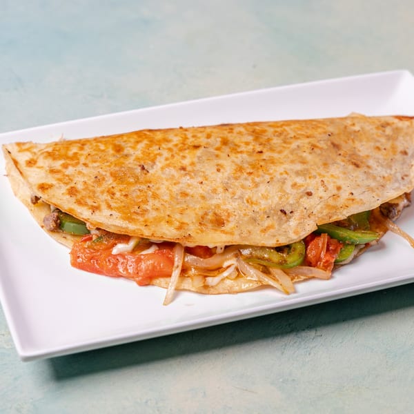 A golden-brown quesadilla filled with meat, melted cheese, green peppers, onions, and tomatoes on a white plate.