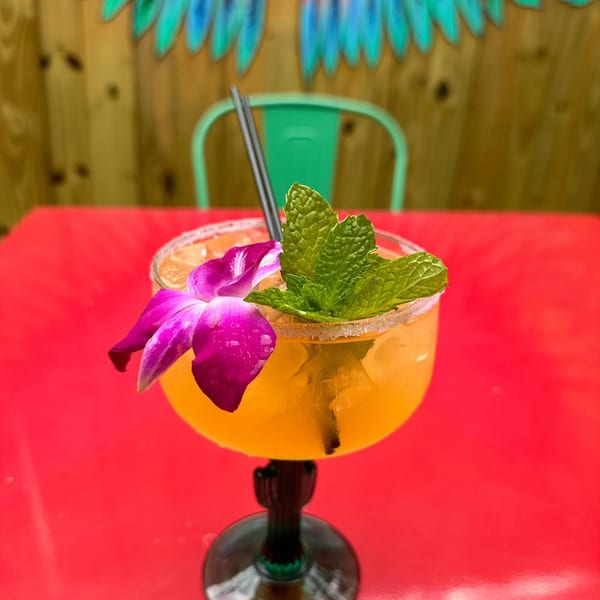 A vibrant orange cocktail with a purple orchid and mint garnish sits on a red table.