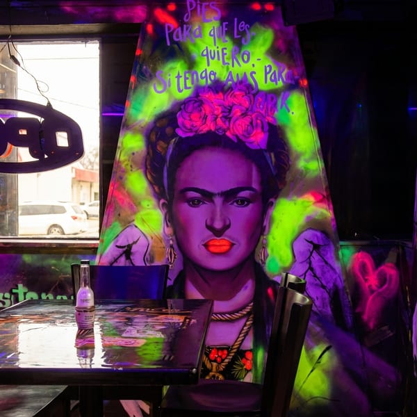 Restaurant interior with neon Frida Kahlo mural, an "OPEN" sign in the window, and a table.
