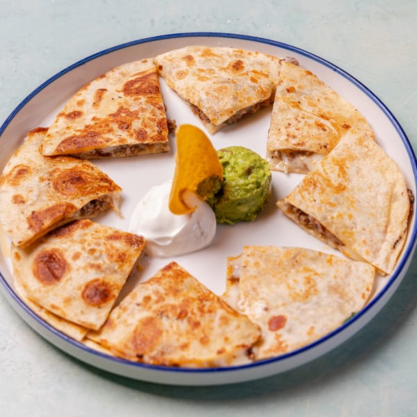 Plate of grilled quesadilla wedges surrounding guacamole, sour cream, and a tortilla chip.