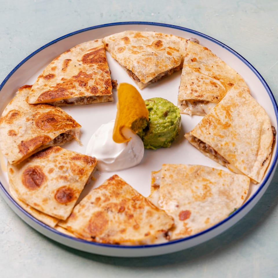 Plate of grilled quesadilla wedges surrounding guacamole, sour cream, and a tortilla chip.