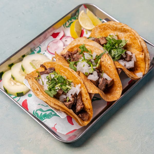 Three carne asada tacos with cilantro and onions, served with lime, radishes, and cucumbers.