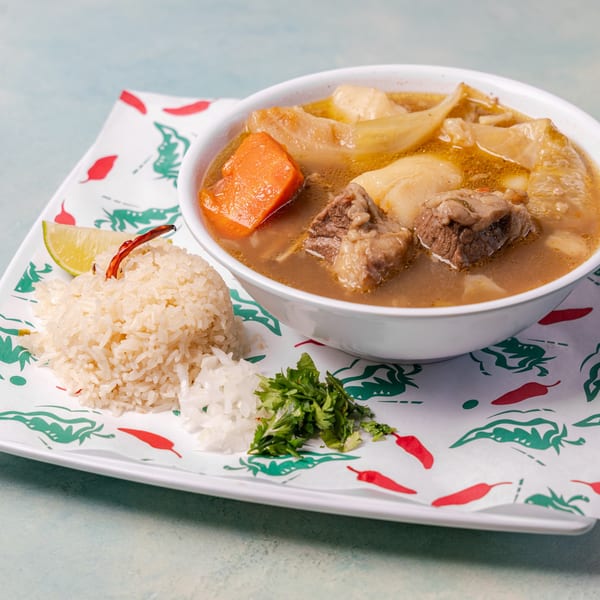 Bowl of beef soup with cabbage and carrots served alongside white rice, lime, chili, and fresh herbs.