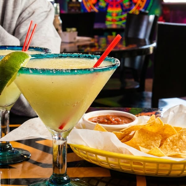 Two margaritas with lime wedges and red straws, alongside a basket of chips and salsa on a dark table.