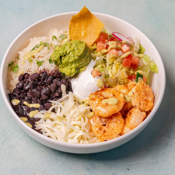 Burrito bowl topped with grilled shrimp, rice, black beans, guacamole, cheese, salad, sour cream, sauce, and tortilla chips.