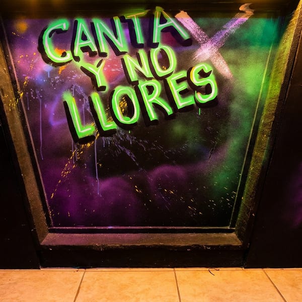 Neon green sign "CANTA Y NO LLORES" on a dark wall with purple and green spray paint.