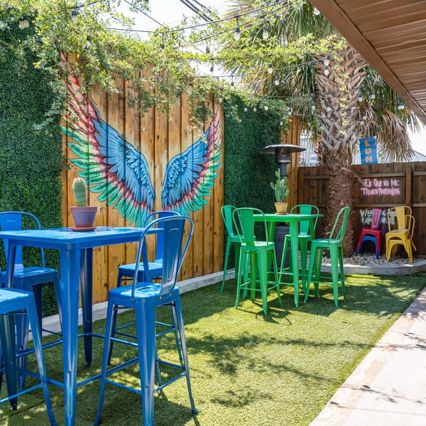 Colorful outdoor patio with blue and green high-top tables, a wing mural, and string lights.