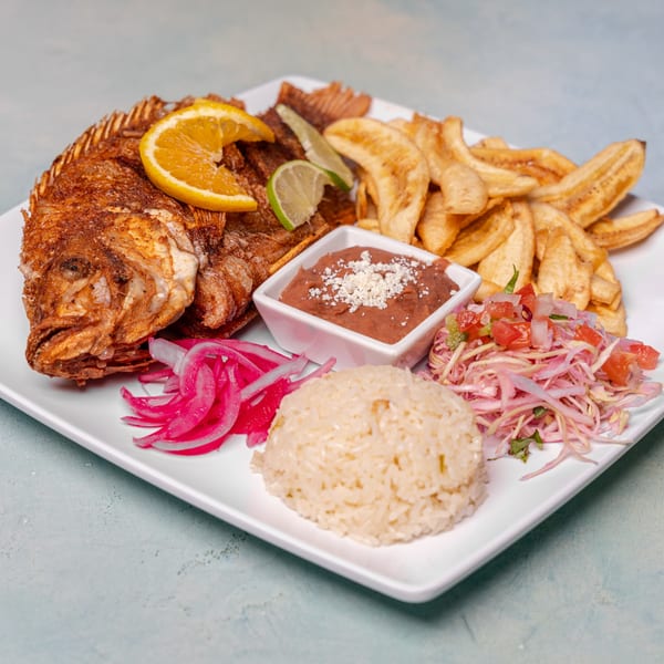 Platter of fried whole fish with rice, beans, plantains, cabbage slaw, pickled onions, and citrus garnishes.
