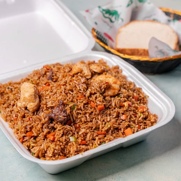 Takeout container filled with seasoned fried rice, chicken, vegetables, and sausage, with sliced bread in a basket behind it.