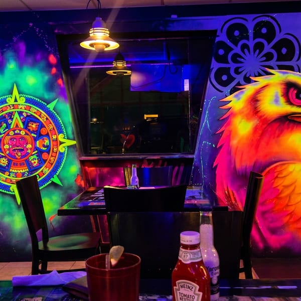 Restaurant interior with neon Aztec calendar and eagle murals, dark tables, and chairs under dim lights.